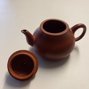 Könnte beinhalten: Kleine, rotbraune Keramik-Teekanne mit passendem Deckel. Die Teekanne hat einen runden Körper, einen gebogenen Griff und einen Ausguss. Der Deckel liegt daneben und gibt die Öffnung frei. Die Teekanne wird wahrscheinlich zum Aufbrühen von Tee verwendet.