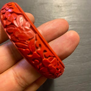 May include: A bright red bangle bracelet featuring detailed floral carvings. The bracelet displays a rich, textured design with relief patterns of flowers and leaves. The vivid red colour and intricate carvings create a striking piece of jewellery, suitable for adding a touch of elegance.