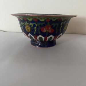 Op de afbeelding: Een decoratieve kom met een uitlopende rand, met een donkerblauwe achtergrond met bloemen- en geometrische patronen in rood, wit, geel en groen. De kom heeft een koperkleurige rand en een voet.