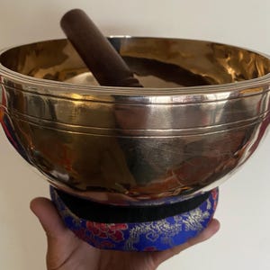May include: A silver singing bowl with a wooden striker, resting on a blue and red patterned cushion. The bowl has a shiny, reflective surface and a dark brown wooden striker. The bowl is made of metal.