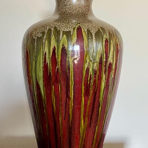 May include: A tall ceramic vase with a flared rim, featuring a glossy finish. The upper neck is speckled brown, transitioning into a vibrant red body with vertical, dripping streaks of lime green glaze that extend downward from the shoulder.