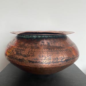 Op de afbeelding: Een gehamerde koperen kom met een brede, licht uitlopende rand. De kom heeft een afgeronde body en een donkere patina rond de rand, met een getextureerd oppervlak. De koperkleur varieert van roodbruin tot een donkerdere, verouderde tint.