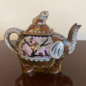 Op de afbeelding: Een theepot in de vorm van een olifant met ingewikkeld cloisonné emailwerk in tinten bruin, roze en goud. Het deksel is bekroond met een klein dierenfiguurtje en het lichaam is versierd met bloemtakmotieven en gedetailleerde patronen.