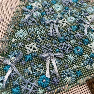 Peut inclure: Gros plan sur un tissu vert avec des broderies bleues et turquoise. La broderie pr&eacute;sente une vari&eacute;t&eacute; de motifs, notamment des n&oelig;uds, des &eacute;toiles et des fleurs.