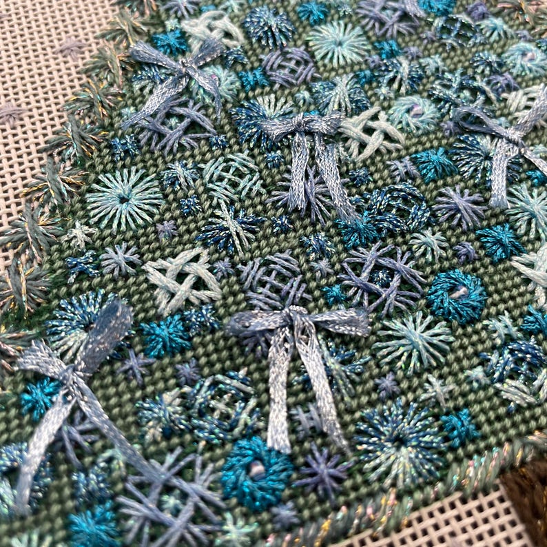 Peut inclure: Gros plan sur un tissu vert avec des broderies bleues et turquoise. La broderie pr&eacute;sente une vari&eacute;t&eacute; de motifs, notamment des n&oelig;uds, des &eacute;toiles et des fleurs.