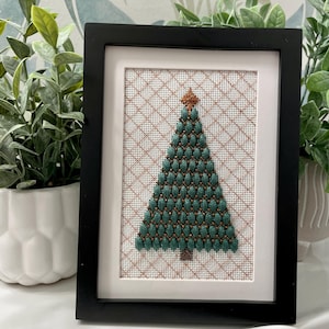 May include: Framed Christmas tree needlepoint art. The tree is green with a copper-coloured star and trunk. The background is a white grid pattern. The artwork is in a black frame.