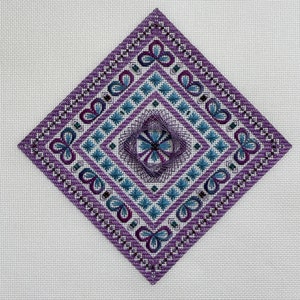以下が含まれることがあります： 白の布地に、紫と青の色合いでダイヤモンド型の刺繍模様が施されています。中央には花柄のモチーフが刺繍されています。