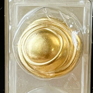 May include: A gold-colored, circular, decorative object with concentric rings. The object is encased in clear plastic.