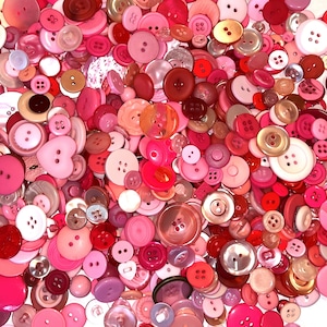 May include: A large collection of pink, red, and white buttons of various sizes and shapes. The buttons are made of plastic, metal, and fabric.