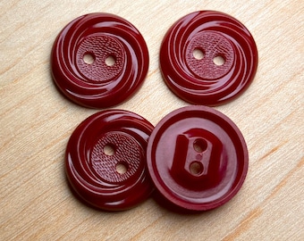 May include: Four burgundy-colored buttons with two holes each. The buttons have a spiral design on the face, with a textured center. The buttons are arranged on a light wood surface.