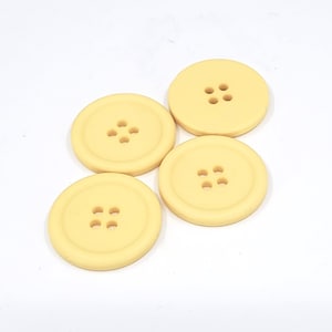 May include: Four large, round, pale yellow buttons with four holes each. The buttons are arranged on a white background. These buttons are suitable for sewing projects, crafts, or adding a decorative touch to clothing.