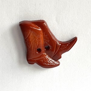 May include: A brown ceramic button shaped like a pair of cowboy boots. The button has two holes for sewing.