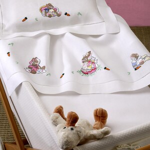 Puede incluir: Juego de ropa de cama para bebé blanco con diseños de conejitos de Pascua y zanahorias bordados. El juego incluye una funda de almohada y una manta, colocados en una cuna de madera. Un peluche de conejito descansa dentro de la cuna.