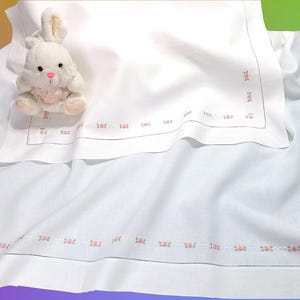 Può includere: Una copertina per neonati bianca con un delicato bordo ricamato rosa. Un giocattolo di peluche di coniglio bianco è seduto sopra la coperta.