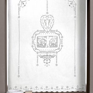Op de afbeelding: Witte raamgordijn met ingewikkelde uitgesneden ontwerpen. Het gordijn heeft een hartvormig ontwerp, bloempatronen en decoratieve randen. De onderkant heeft een geschulpte rand. Het gordijn hangt in een raamkozijn.