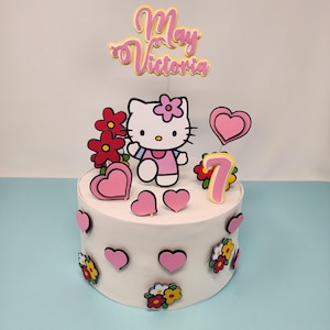 May include: A white cake decorated with a Hello Kitty character, pink flowers, pink hearts, and a pink number 7. The cake also has a pink and yellow topper that says "May Victoria".
