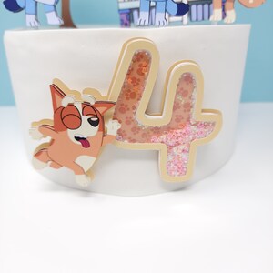 May include: A white cake with a brown and white cartoon dog and a gold glitter number four on top. The number four has a pink and orange glitter inside.