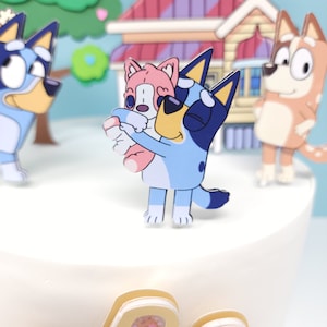 May include: A blue and white cartoon dog holding a pink and yellow cartoon puppy. The dog is wearing sunglasses and has a blue and white patterned tail. The puppy has a pink nose and a yellow belly. The figures are on a white cake.