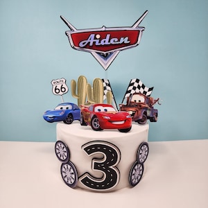 Op de afbeelding: Een witte taart versierd met Cars-thema toppers, waaronder Bliksem McQueen, Takel en een Route 66 bord. De taart heeft een groot nummer 3 en auto wiel accenten, met een banner met de tekst "Aiden".