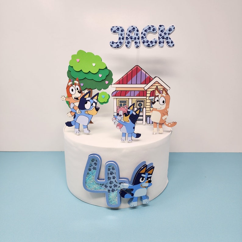 May include: A blue and white cake topper set featuring characters from the cartoon show Bluey. The set includes a house, a tree, a number 4, and the characters Bluey, Bingo, and Bandit. The topper also includes the name "Jack" in blue and white letters.
