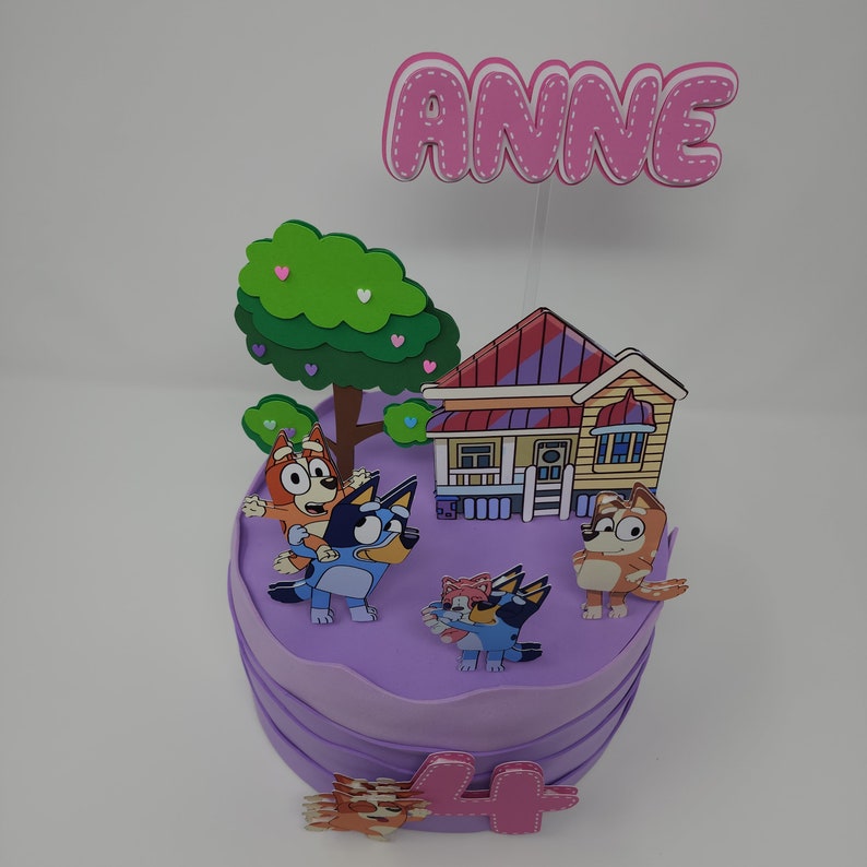 Puede incluir: Pastel morado decorado con una casa, un &aacute;rbol y personajes del programa de dibujos animados "Bluey". El pastel est&aacute; cubierto con un letrero rosa que dice "ANNE" y un n&uacute;mero rosa "4".