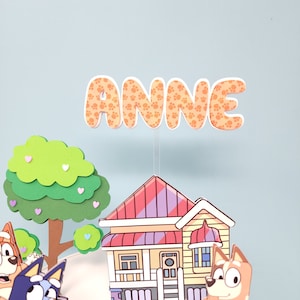 May include: A colorful cake topper featuring a house, a tree, and two cartoon dogs. The topper also includes the name "ANNE" in orange letters with paw print designs.