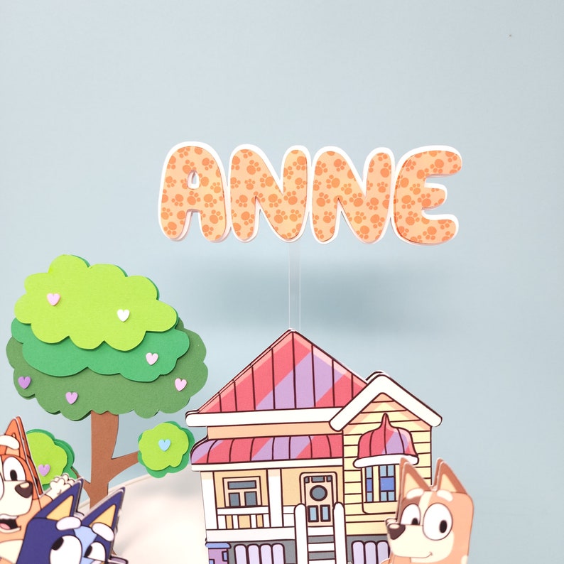 May include: A colorful cake topper featuring a house, a tree, and two cartoon dogs. The topper also includes the name "ANNE" in orange letters with paw print designs.
