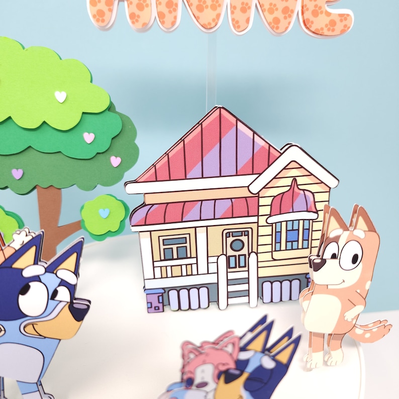 May include: A colorful papercraft scene featuring a yellow house with a red and purple striped roof, a green tree, and several cartoon dogs. The dogs are blue, brown, and pink, and they are all wearing different expressions.