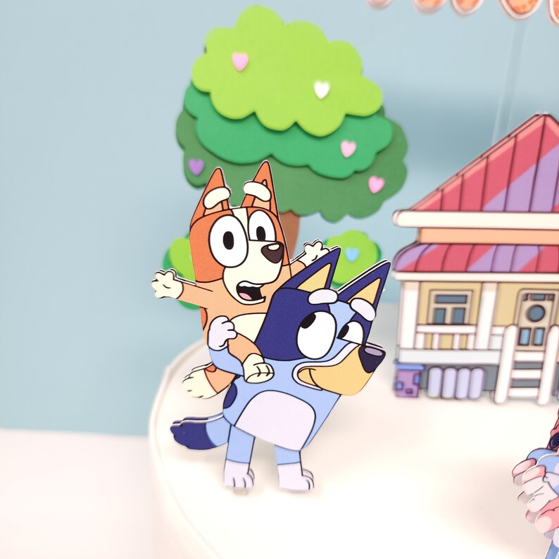 May include: A blue and white cartoon dog with a brown and orange cartoon dog riding on its back. The dogs are standing on a white cake with a green tree and a red and white house in the background.