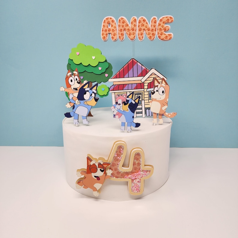 May include: A white cake decorated with a '4' in gold glitter, a house, a tree, and characters from the cartoon 'Bluey'. The cake also has the name 'Anne' on top.
