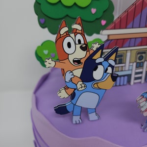 Puede incluir: Un pastel morado decorado con personajes de dibujos animados del programa Bluey. Los personajes son un perro azul y un perro naranja. El perro azul est&aacute; de pie sobre el pastel y el perro naranja est&aacute; sentado en la espalda del perro azul. El pastel est&aacute; decorado con un &aacute;rbol verde y una casa rosa.