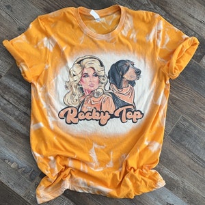 Peut inclure: Un t-shirt orange vif &agrave; manches courtes avec un effet tie-dye d&eacute;color&eacute;. Le motif central repr&eacute;sente une femme blonde et un chien de chasse noir et feu, avec le texte "Rocky Top" &eacute;crit dans une police orange arrondie sous les personnages.