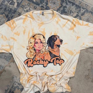 Peut inclure: Un t-shirt &agrave; manches courtes avec un motif tie-dye cr&egrave;me et orange. Le graphique central repr&eacute;sente une femme blonde et un chien de chasse noir et feu portant un bandana. Sous les personnages, le texte Rocky Top est imprim&eacute; en orange.