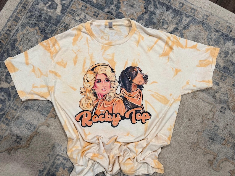 Peut inclure: Un t-shirt &agrave; manches courtes avec un motif tie-dye cr&egrave;me et orange. Le graphique central repr&eacute;sente une femme blonde et un chien de chasse noir et feu portant un bandana. Sous les personnages, le texte Rocky Top est imprim&eacute; en orange.