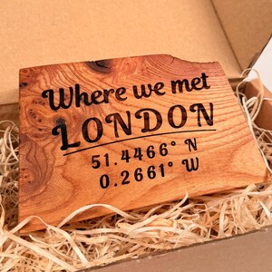 May include: A wooden plaque with the text "Where we met LONDON" and coordinates 51.4466° N 0.2661° W, presented in a cardboard box with straw. The plaque has a natural wood grain.