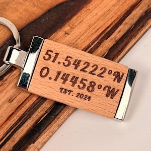 May include: A wooden rectangular keyring with engraved coordinates and the text "EST. 2024". The keyring has silver-coloured metal accents and a silver key ring. The coordinates are 51.54222°N and 0.14458°W.