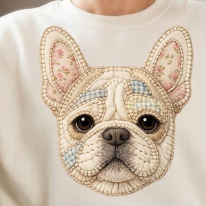 Puede incluir: Sudadera de color crema con una ilustración detallada de la cara de un bulldog francés. Las orejas y la frente del perro tienen patrones florales y a cuadros. Los ojos del perro son oscuros y la nariz es negra.