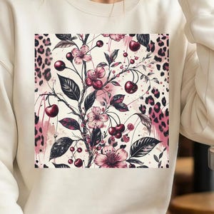 Op de afbeelding: Crèmekleurige sweatshirt met een vierkante afbeelding van een bloemen- en kersendesign. De afbeelding bevat roze bloemen, donkere kersen en zwarte bladeren tegen een crèmekleurige achtergrond met luipaardprint accenten. De sweatshirt heeft lange mouwen.