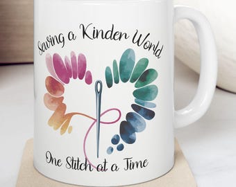 May include: White ceramic mug with a heart-shaped design featuring a needle and thread, and the text "Saving a Kinder World One Stitch at a Time." The heart is composed of watercolor-style segments in various colors.