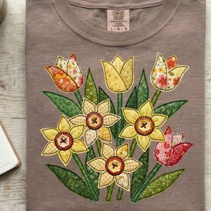 May include: A taupe t-shirt with an embroidered floral design. The design features yellow daffodils and tulips with green leaves. The flowers have patterned details in yellow, orange, and pink. The shirt has a crew neck and short sleeves.