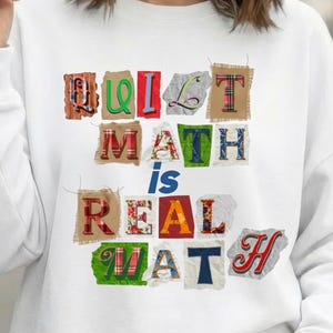 May include: White sweatshirt with the text "QUIT MATH is REAL MATH" in colorful, patchwork-style letters. The letters have various patterns and textures, including plaid, floral, and burlap.