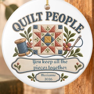 May include: A round white ceramic ornament featuring the text QUILT PEOPLE at the top, a central quilt block graphic, a blue thread spool, a needle, and a banner with the text You keep all the pieces together, Marianne, and 2026.
