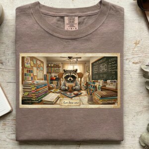 Puede incluir: Camiseta de color malva con un estampado rectangular de un mapache en un aula, bebiendo de una taza. El gráfico incluye libros, una pizarra y el texto "i live here now". La camiseta tiene cuello redondo y mangas cortas.