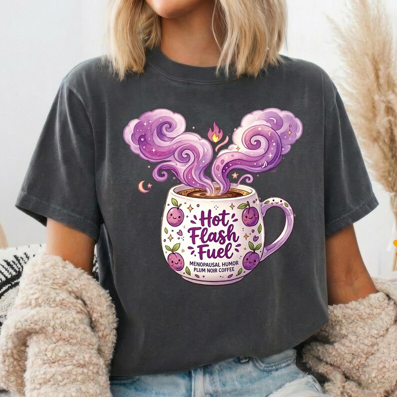 Puede incluir: Una camiseta gris carb&oacute;n con un gr&aacute;fico de una taza de caf&eacute; blanca con vapor morado. La taza muestra el texto Hot Flash Fuel, Menopausal Humor, Plum Noir Coffee, rodeado de peque&ntilde;as ilustraciones de ciruelas moradas y estrellas.