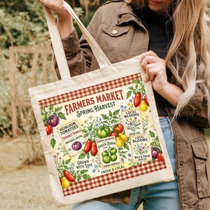 Puede incluir: Bolso tote color crema con un diseño de "Farmers Market Spring Harvest". El diseño incluye ilustraciones de tomates, flores y texto. El bolso tiene un borde a cuadros rojo y blanco y asas color crema.