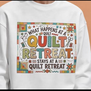 May include: White sweatshirt with a colorful graphic that reads "WHAT HAPPENS AT A QUILT RETREAT STAYS AT A QUILT RETREAT." The design includes images of sewing tools, stars, and quilt block patterns. The sweatshirt is a crew neck style.