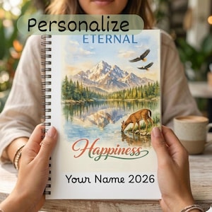 May include: Spiral-bound notebook with a scenic landscape illustration. The artwork depicts a mountain range reflected in a lake, with a deer drinking water and two eagles soaring above. The notebook has the words "Personalize", "Eternal", and "Happiness" with the year 2026.