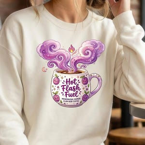 Puede incluir: Sudadera color crema con una ilustraci&oacute;n caprichosa de una taza de caf&eacute;. La taza est&aacute; adornada con ciruelas moradas y las palabras "Hot Flash Fuel". Vapor morado se eleva del caf&eacute;, con el texto "Menopausal Humor Plum Noir Coffee" debajo.