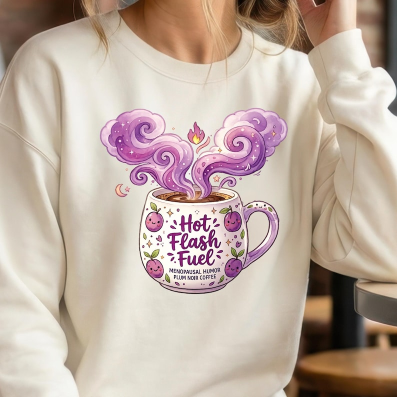 Puede incluir: Sudadera color crema con una ilustraci&oacute;n caprichosa de una taza de caf&eacute;. La taza est&aacute; adornada con ciruelas moradas y las palabras "Hot Flash Fuel". Vapor morado se eleva del caf&eacute;, con el texto "Menopausal Humor Plum Noir Coffee" debajo.