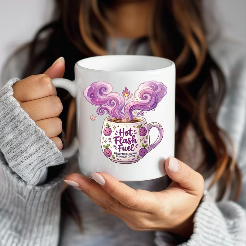 Puede incluir: Taza de caf&eacute; de cer&aacute;mica blanca con el texto "Hot Flash Fuel" y una ilustraci&oacute;n caprichosa de una taza de caf&eacute; humeante con vapor p&uacute;rpura. La taza tambi&eacute;n presenta detalles de ciruelas y hojas, y las palabras "Menopausal Humor Plum Noir Coffee".